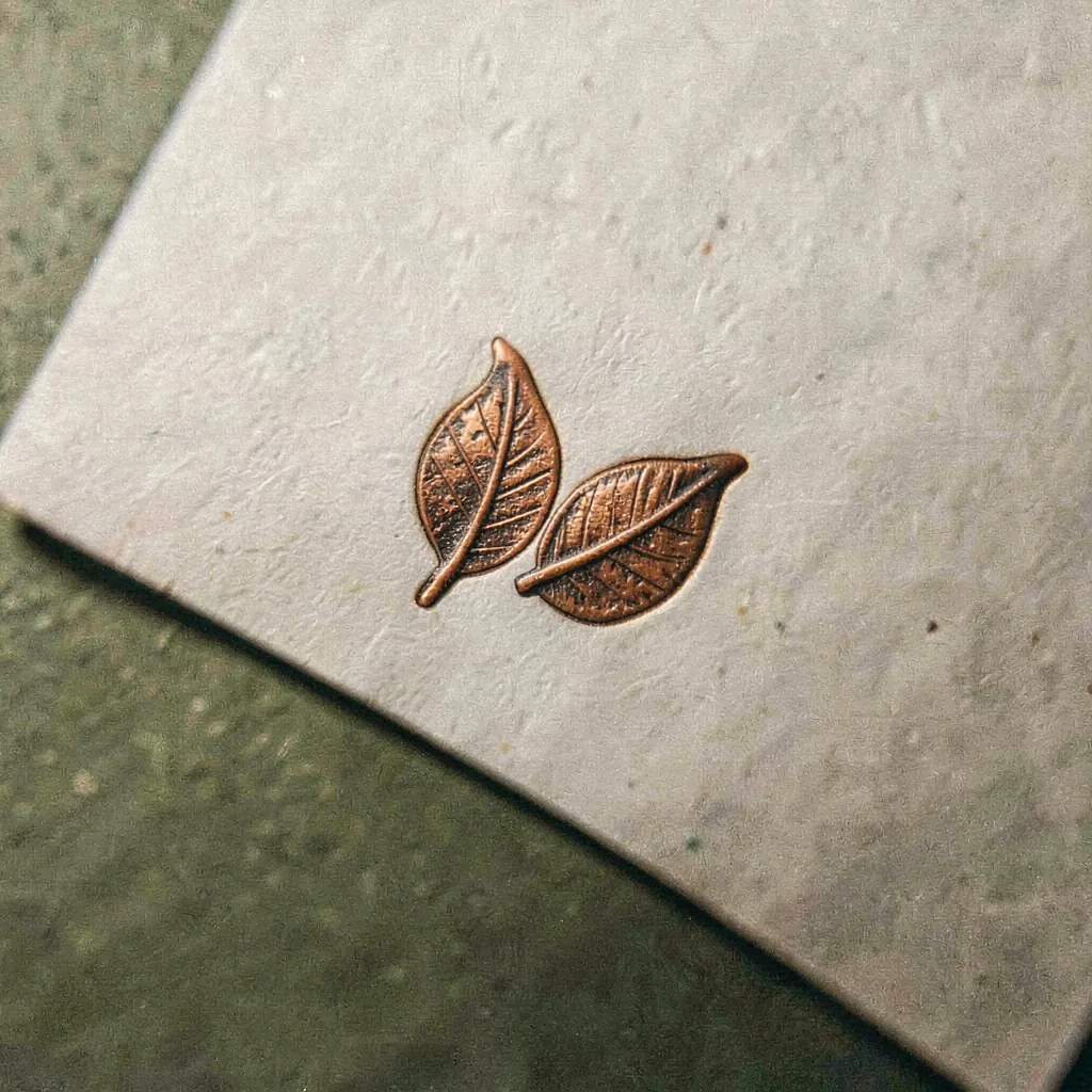 A small copper pressmark — two folded leaves over the studio initials